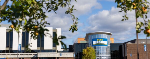 A tree with apples in the foreground frames the SUNY Niagara campus building under a partly cloudy sky.