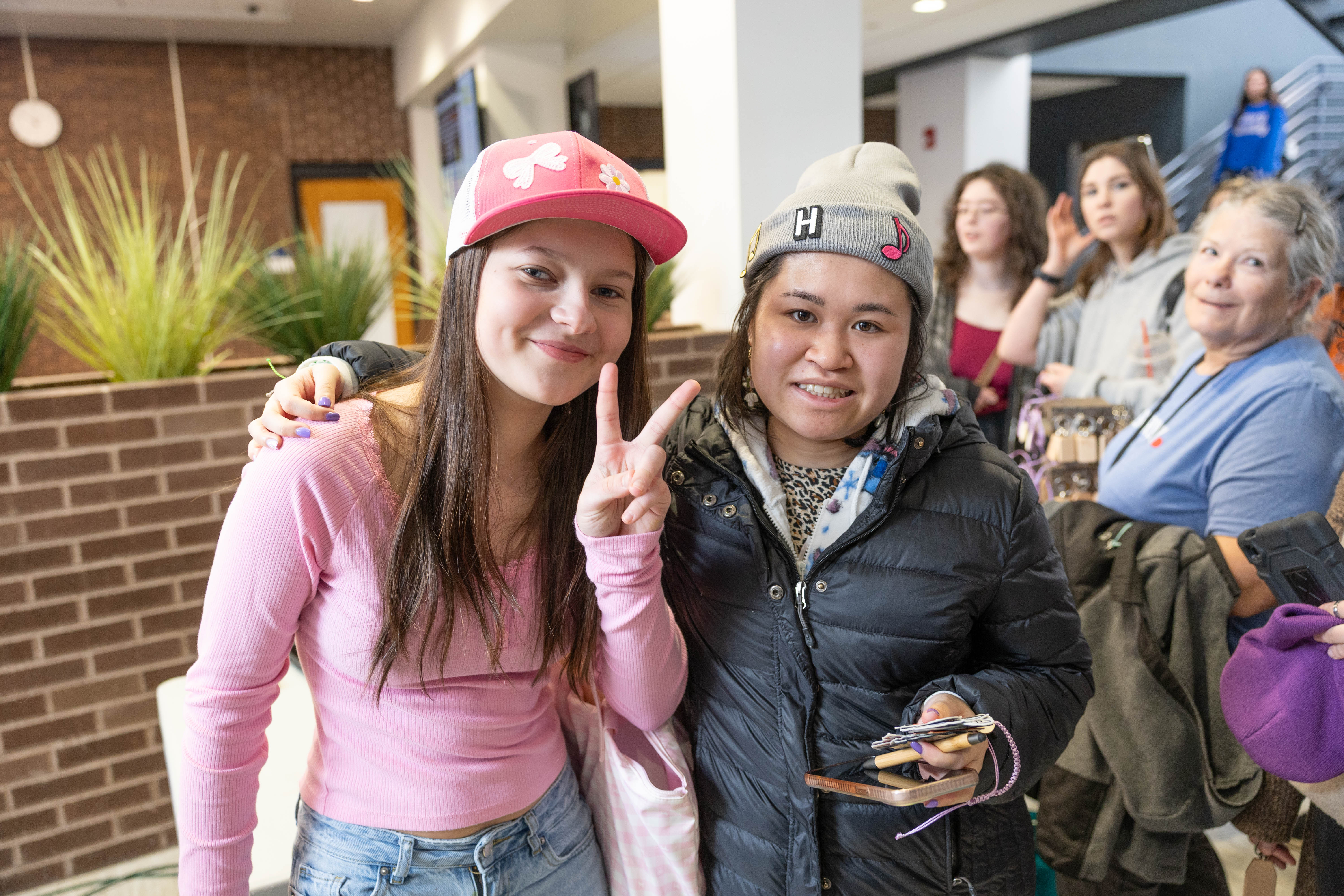 Two women pose and smile indoors; one wears a pink hat and shirt, making a peace sign, while the other wears a black jacket and gray hat. People stand and watch in the background.