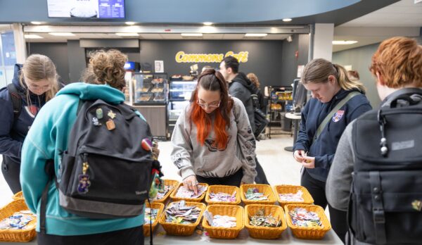 Several people stand around a table with baskets of colorful patches in a cafe setting. A digital menu is visible in the background, and everyone appears to be browsing or selecting items.