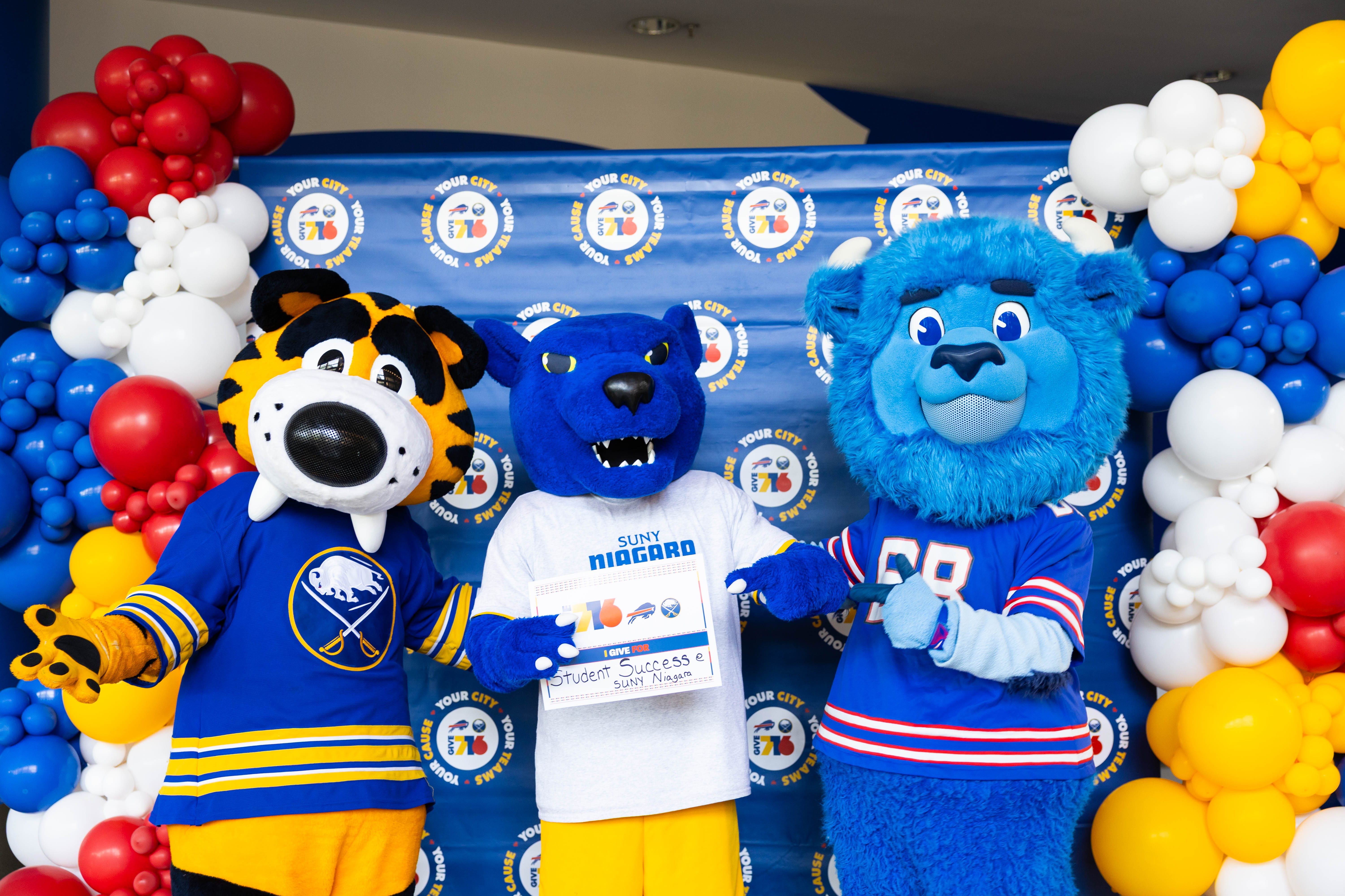 Three colorful sports mascots stand in front of a backdrop with logos and balloon decorations. The center mascot holds a sign. All are smiling and wearing themed costumes and jerseys.