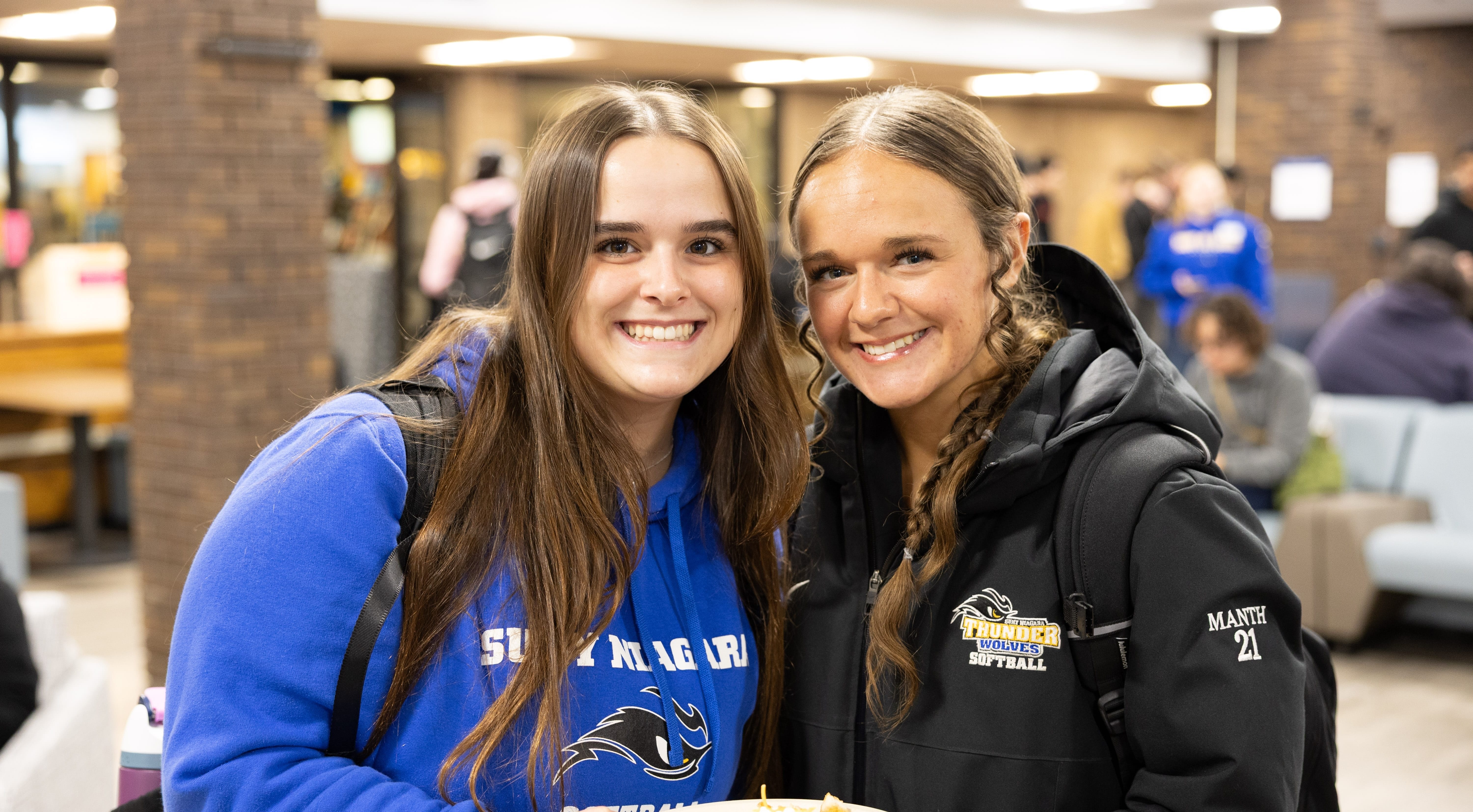 Two young women smile indoors. One holds a plate of food. Both wear blue sports hoodies and backpacks. Other people and lounge furniture are visible in the background.