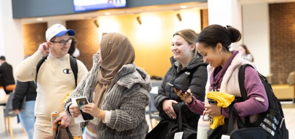 Four people stand around a table with assorted cookies, smiling and looking at their phones. They carry bags and wear casual clothing, appearing cheerful in a bright indoor setting with others in the background.