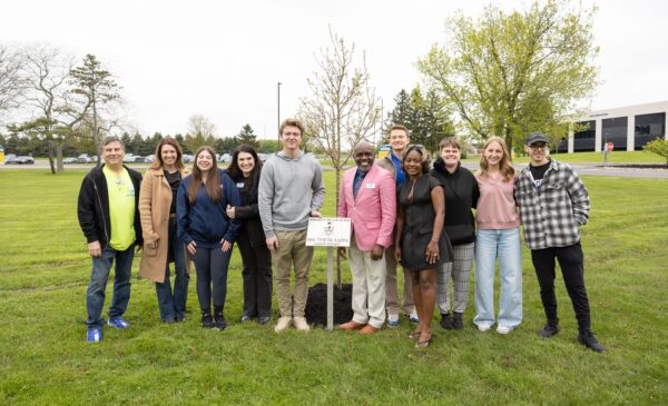 A group of eleven people stand on grass, smiling around a young tree they planted, with a small sign in front. Office buildings and trees are visible in the background.