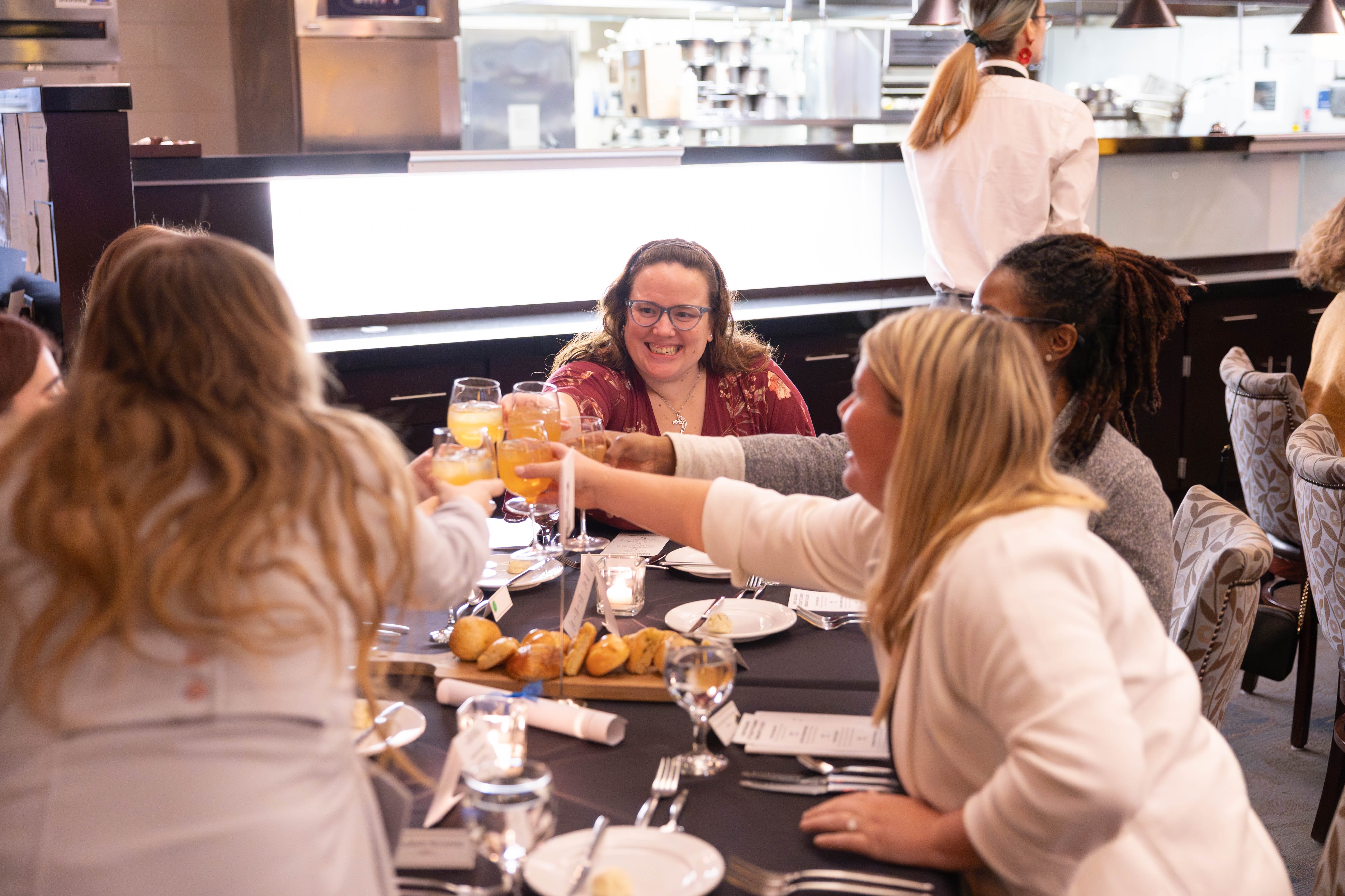 A group of women sit around a restaurant table, smiling and raising glasses for a toast. Food, bread, and candles are on the table, and a chef stands in the background near the kitchen.