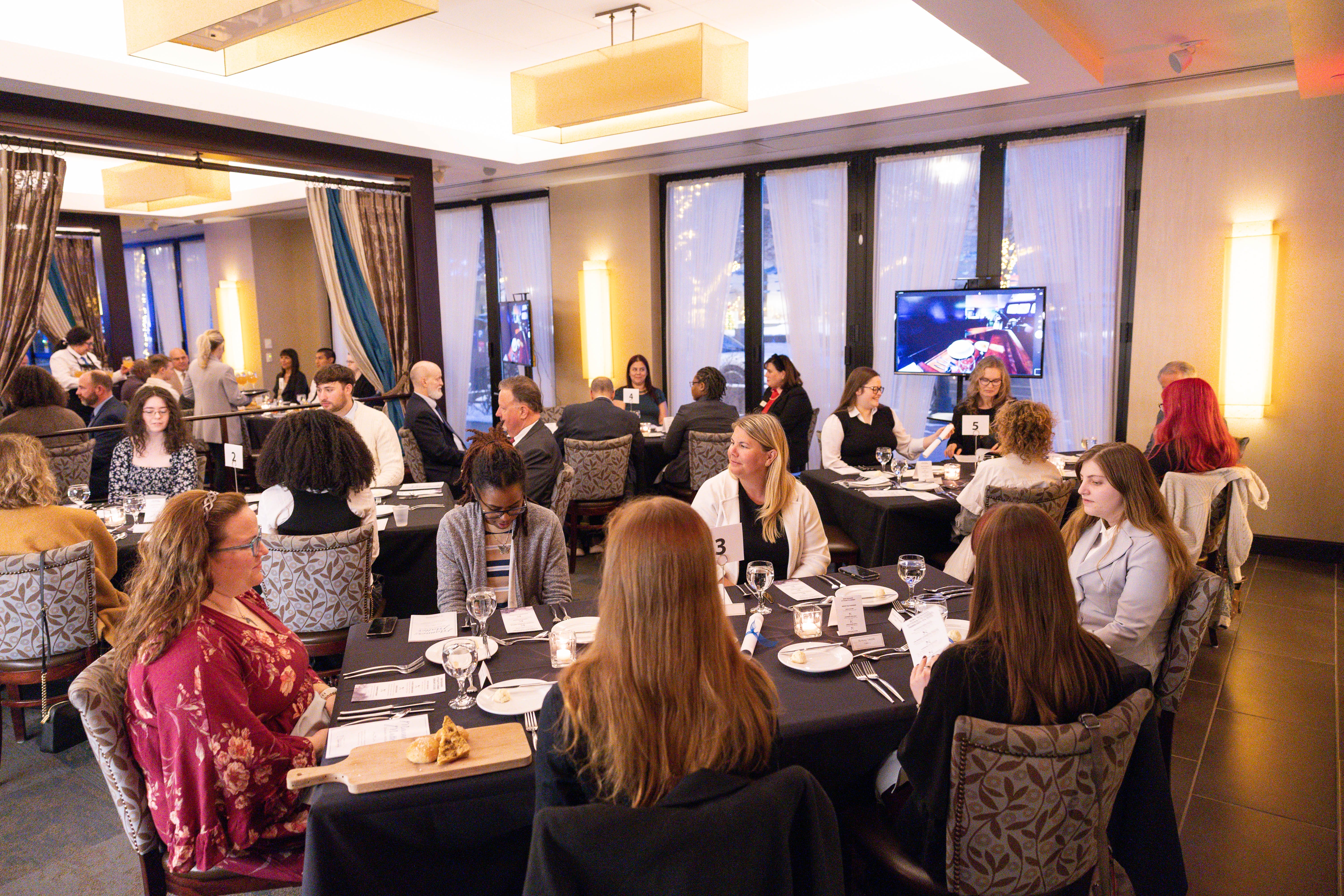 People are seated around multiple tables in a modern, well-lit restaurant, attending a formal event or dinner. Most are engaged in conversation, and some are eating. There are table numbers and a screen visible in the background.