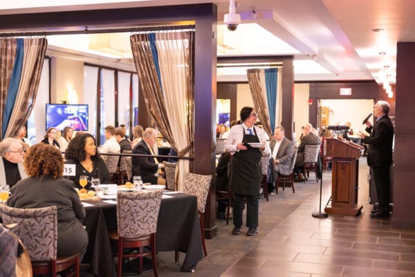 A formal event or conference is taking place in a restaurant, with people seated at tables, a waiter serving, and a person speaking at a podium at the front of the room.