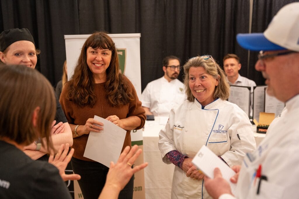 A group of people, including chefs in white uniforms and others holding papers, stand in a circle smiling and talking at an indoor event with a table and banner in the background.