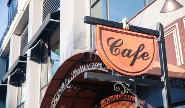 A red and black sign reading Cafe hangs outside a building with large windows and a curved awning, suggesting a welcoming entrance to a coffee shop or café on a sunny day.