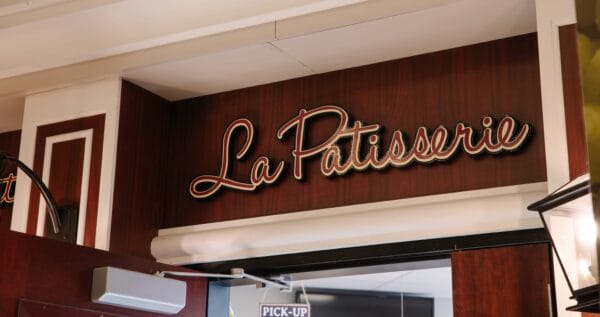 A sign reading La Patisserie in elegant script is mounted on a dark wood-paneled wall above a shop entrance.