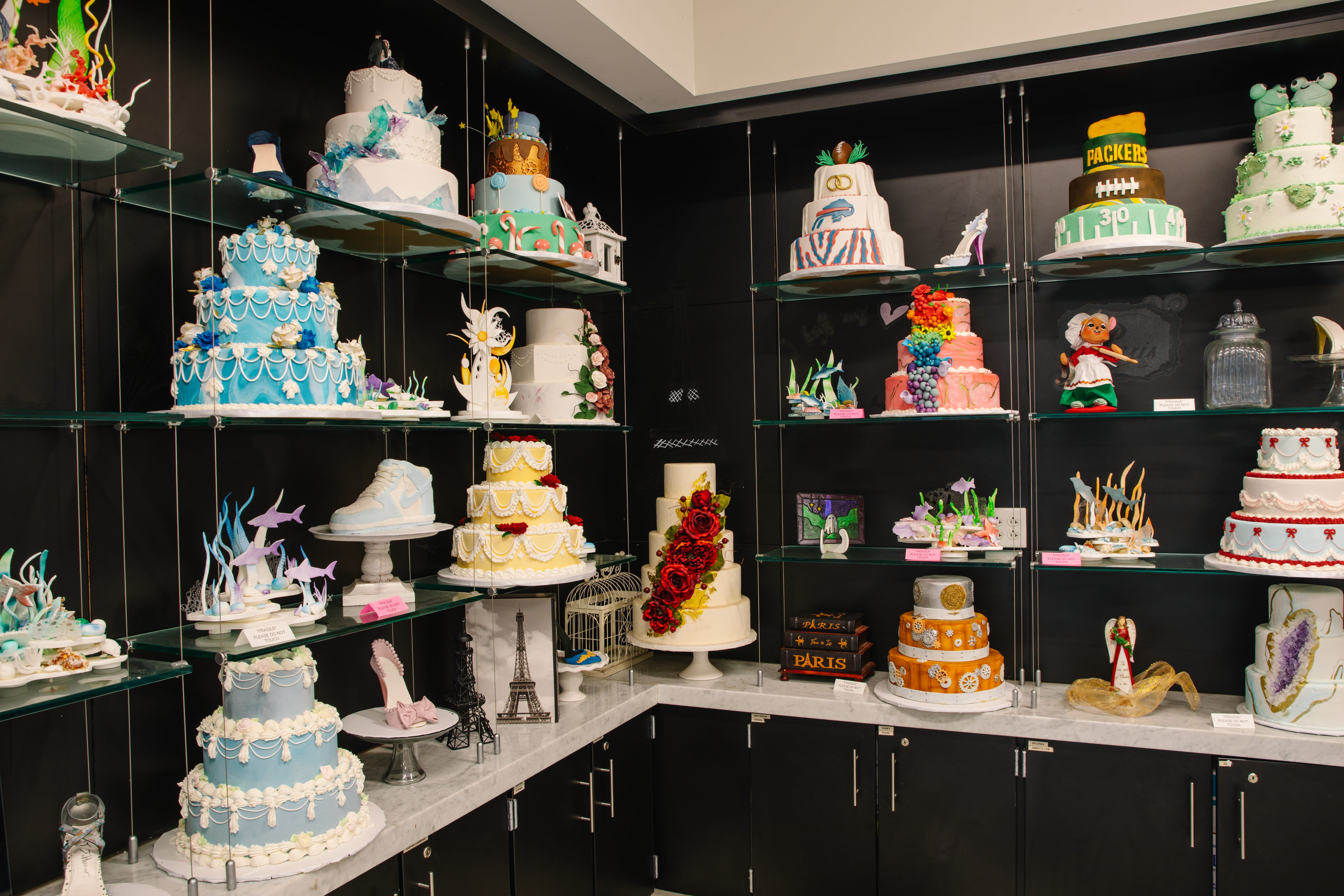 A display of intricately decorated cakes of various shapes, themes, and colors arranged on glass shelves and a marble counter against a black wall. Designs include sports, fashion, flowers, and landmarks.