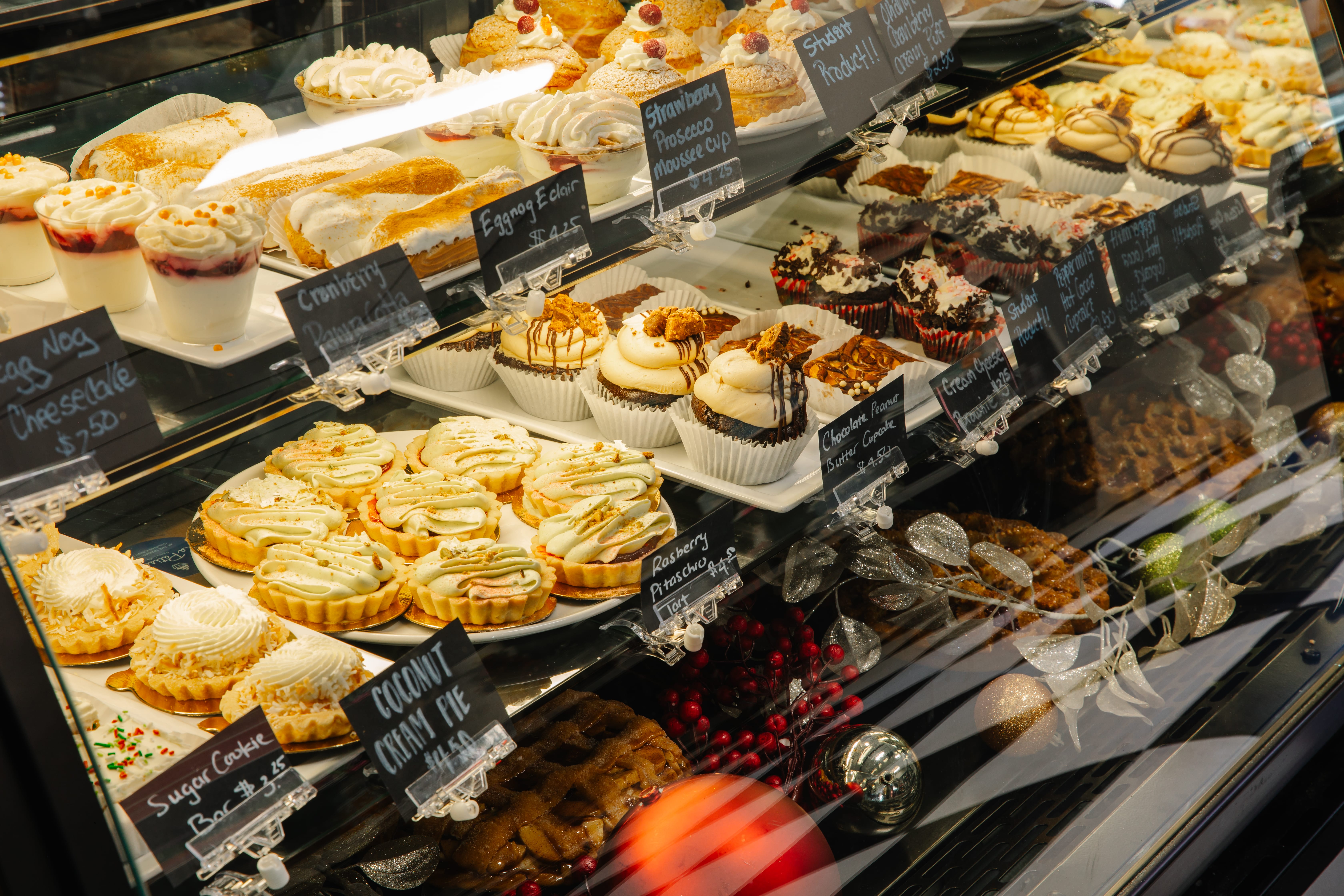 A bakery display case filled with an assortment of cupcakes, tarts, pies, and pastries, each labeled with handwritten signs. The treats are decorated with icing, chocolate, and toppings.