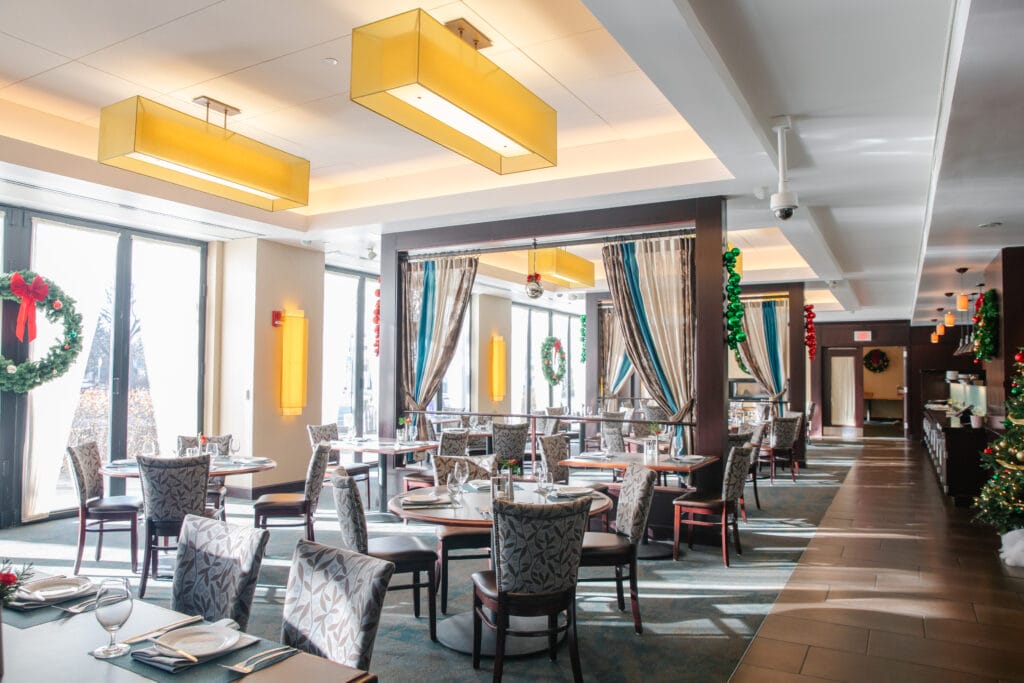 A bright, modern restaurant dining room with tables set for guests. Large windows let in natural light, and holiday wreaths and garlands decorate the space. Neutral tones and patterned chairs create an inviting atmosphere.