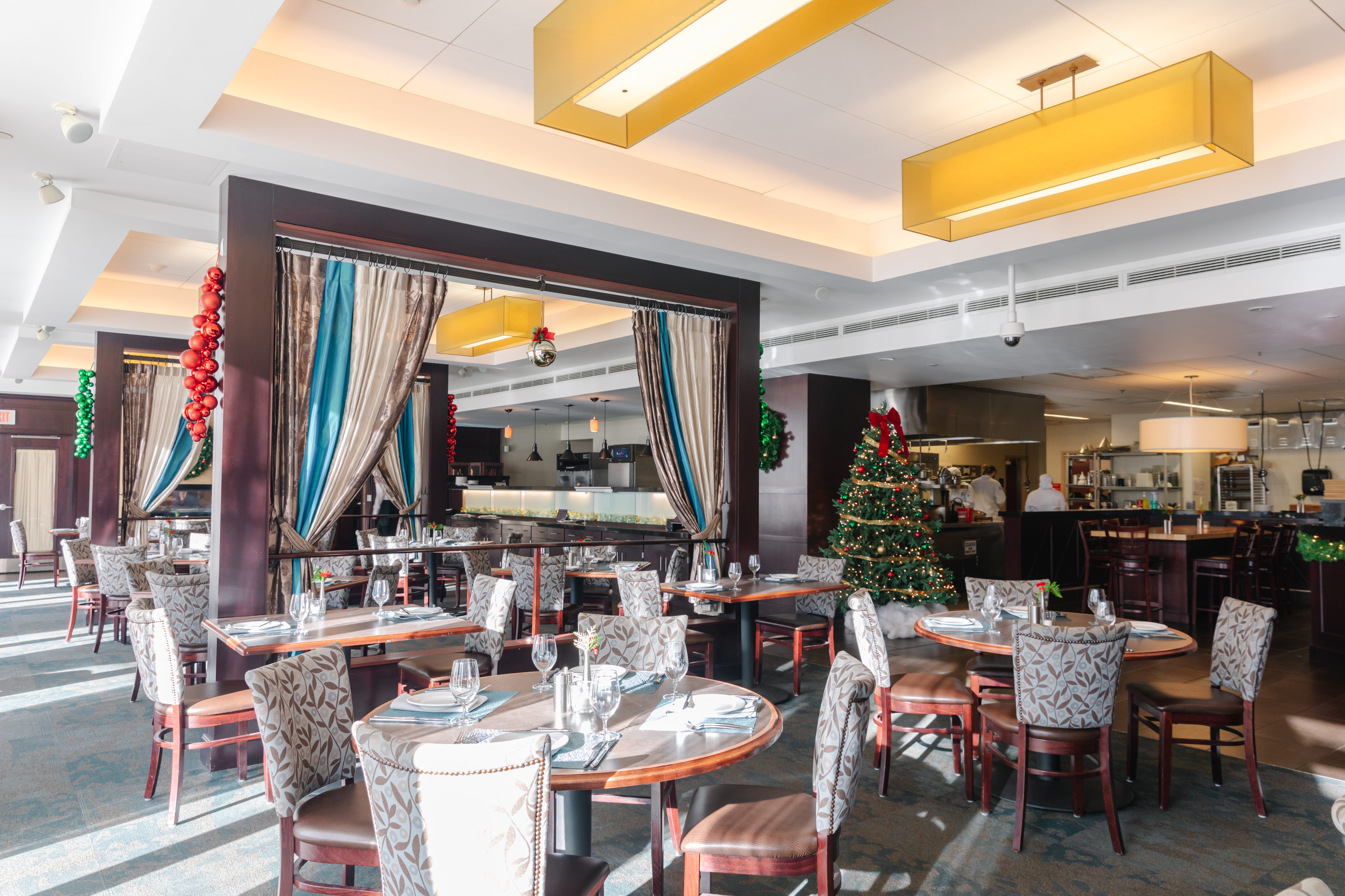 A bright restaurant dining area decorated for Christmas, featuring set tables, patterned chairs, a Christmas tree with ornaments, festive wreaths, and garlands near the kitchen counter.