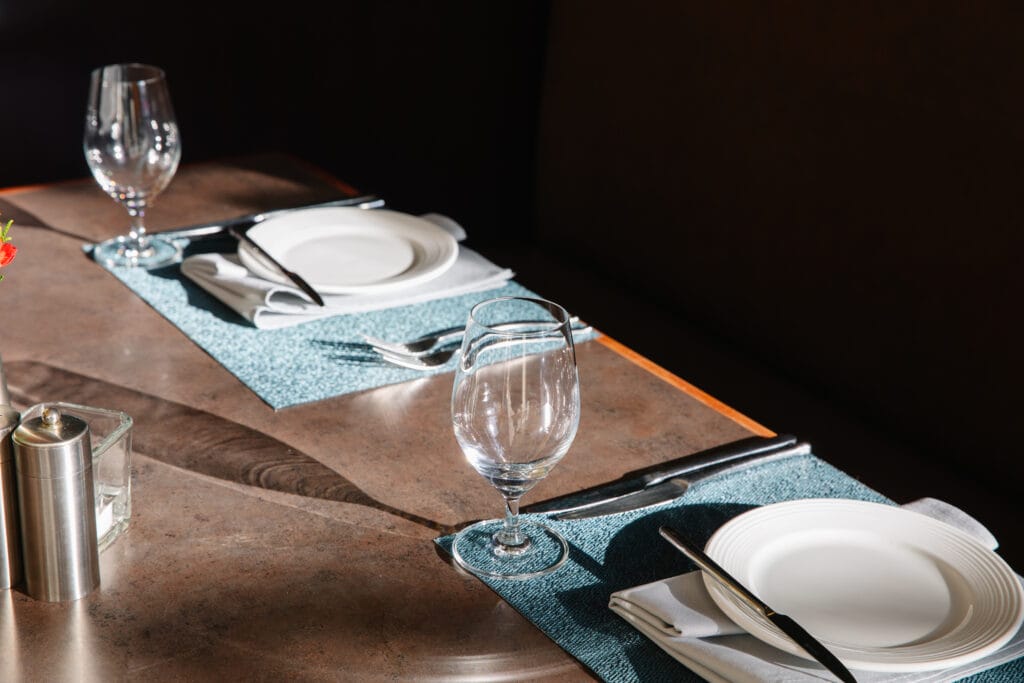 A close-up of a restaurant table set for two with white plates, silverware, and water glasses on blue placemats, lit by sunlight. A salt shaker and a votive candle are also visible on the table.