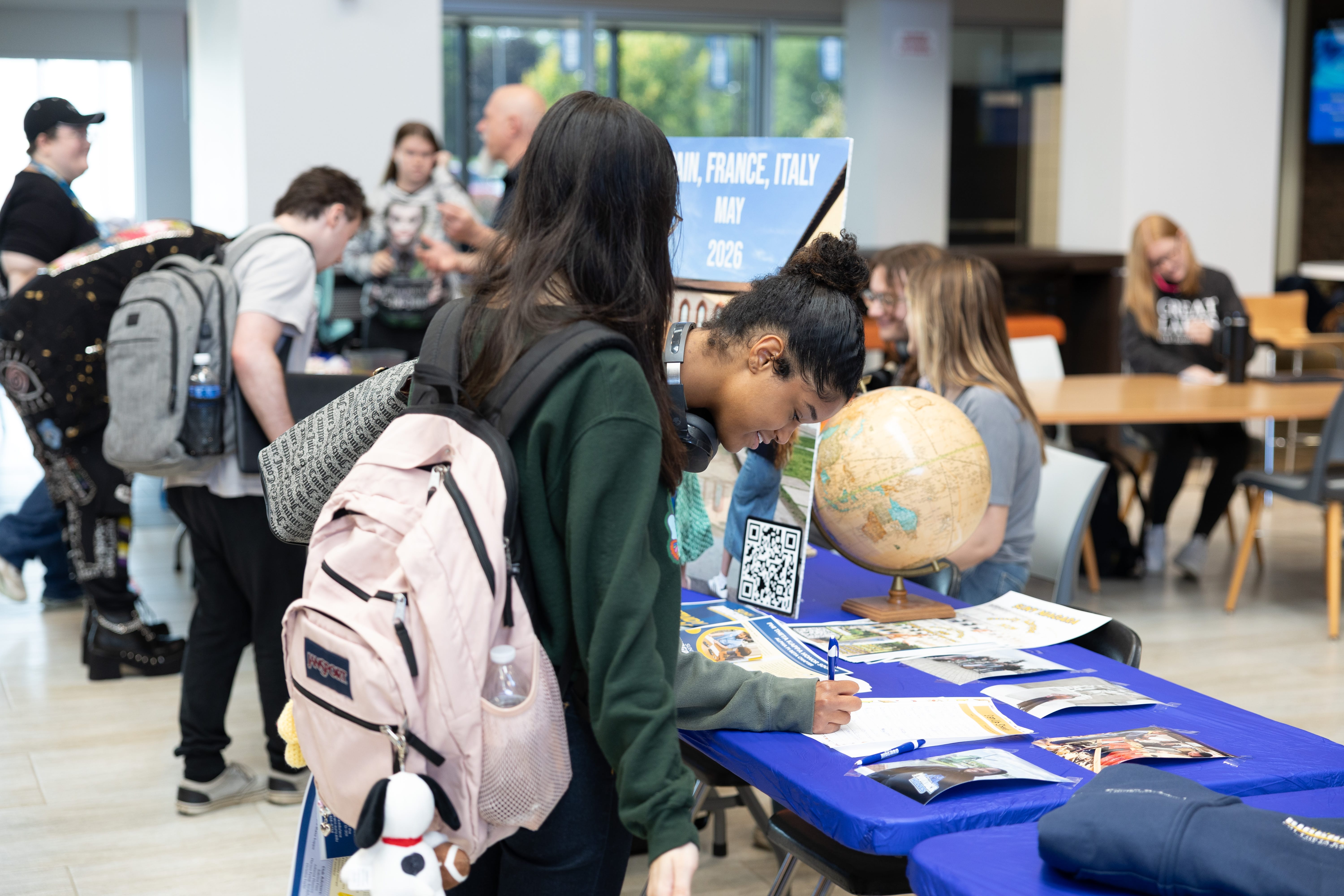 Students gather around a table with travel brochures, a globe, and a sign reading “Spain, France, Italy May 2023.” One student writes on a form while others look at materials or talk nearby in a bright indoor space.