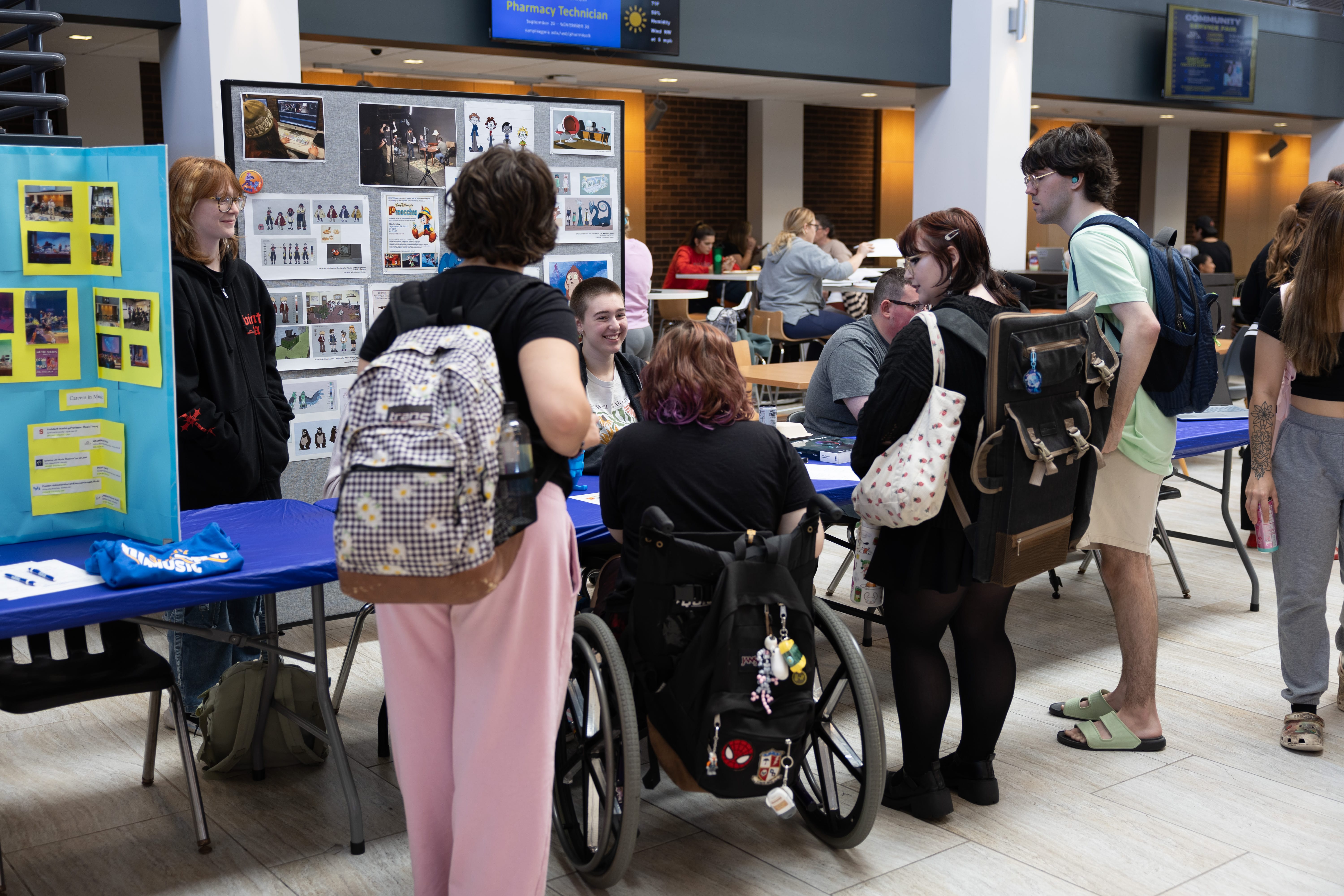 Students gather around informational tables at a school event. Some stand, one uses a wheelchair, and posters are displayed on tables. People are talking and engaging with materials in a bright, indoor common area.