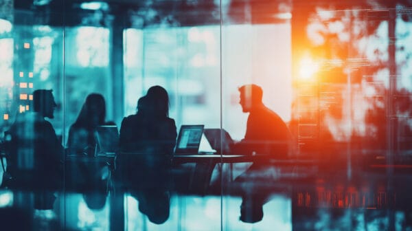 Silhouettes of four people sit around a conference table with laptops in a modern office, sunlight streaming through large windows, creating a vibrant, collaborative atmosphere.
