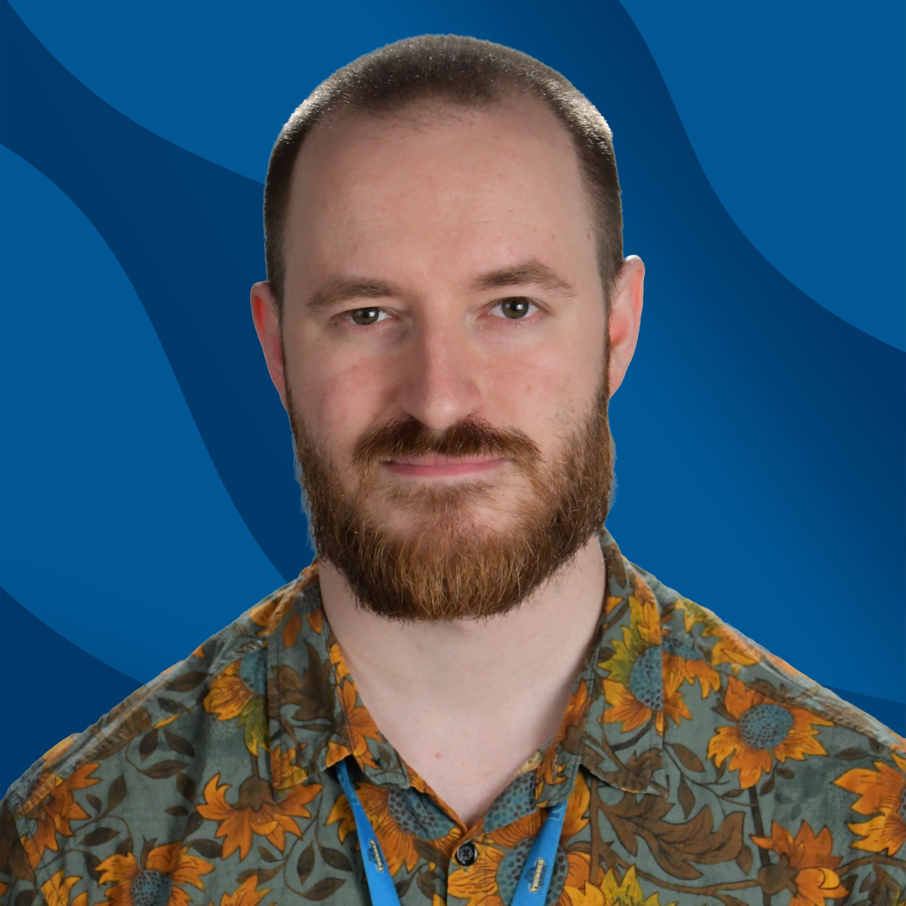 A bearded man with short hair wearing a floral shirt and a lanyard stands in front of a blue, wavy-patterned background.