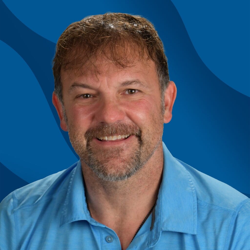 A smiling man with short brown hair and a beard, wearing a light blue collared shirt, poses in front of a blue wavy background.