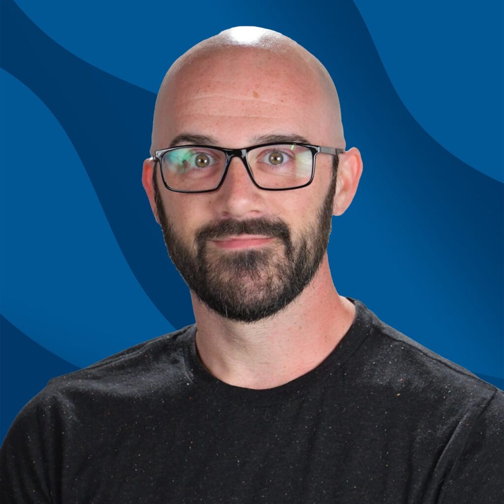 Bald man with black glasses and a beard, wearing a black shirt, poses in front of a blue wavy background.