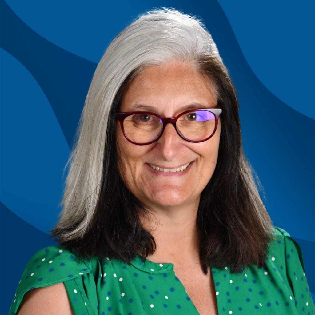 Smiling woman with long black and white hair, wearing glasses and a green top with white dots, in front of a blue abstract background.