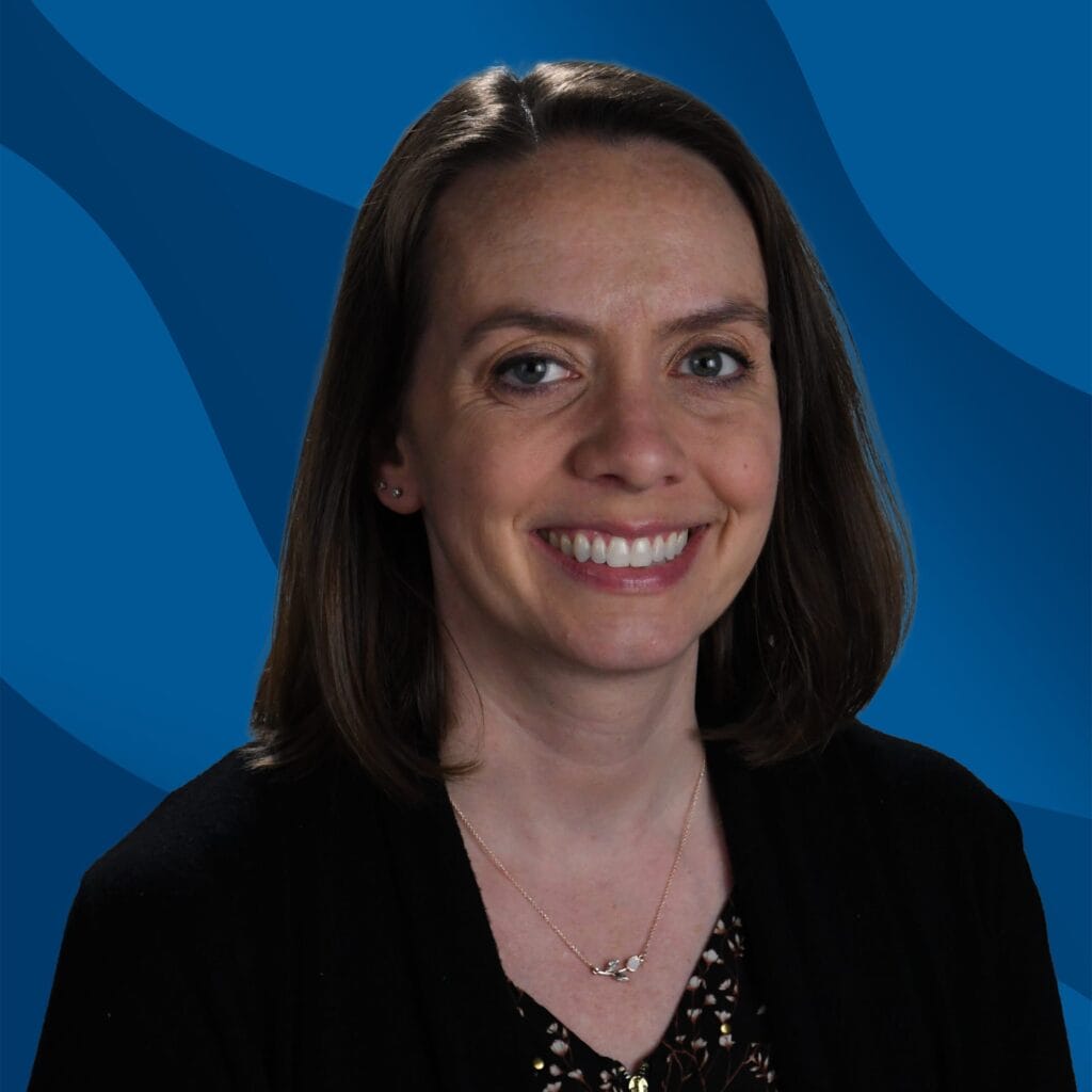 Woman with shoulder-length brown hair smiles at the camera. She is wearing a black top and necklace, with a blue background that has wave-like patterns.
