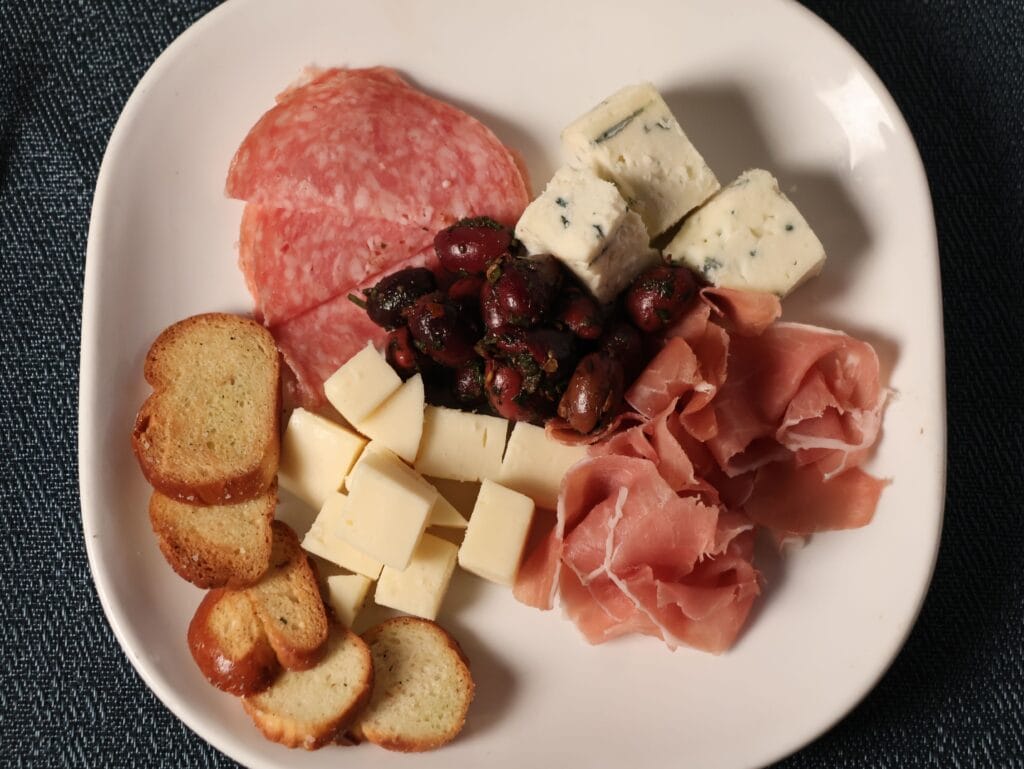 A white plate with sliced salami, prosciutto, cubed cheese, blue cheese, marinated olives, and toasted bread slices, arranged on a blue tablecloth.