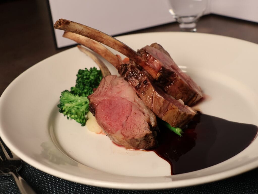 A plate with two racks of lamb, cooked medium rare, served with broccoli, mashed potatoes, and a dark wine sauce.
