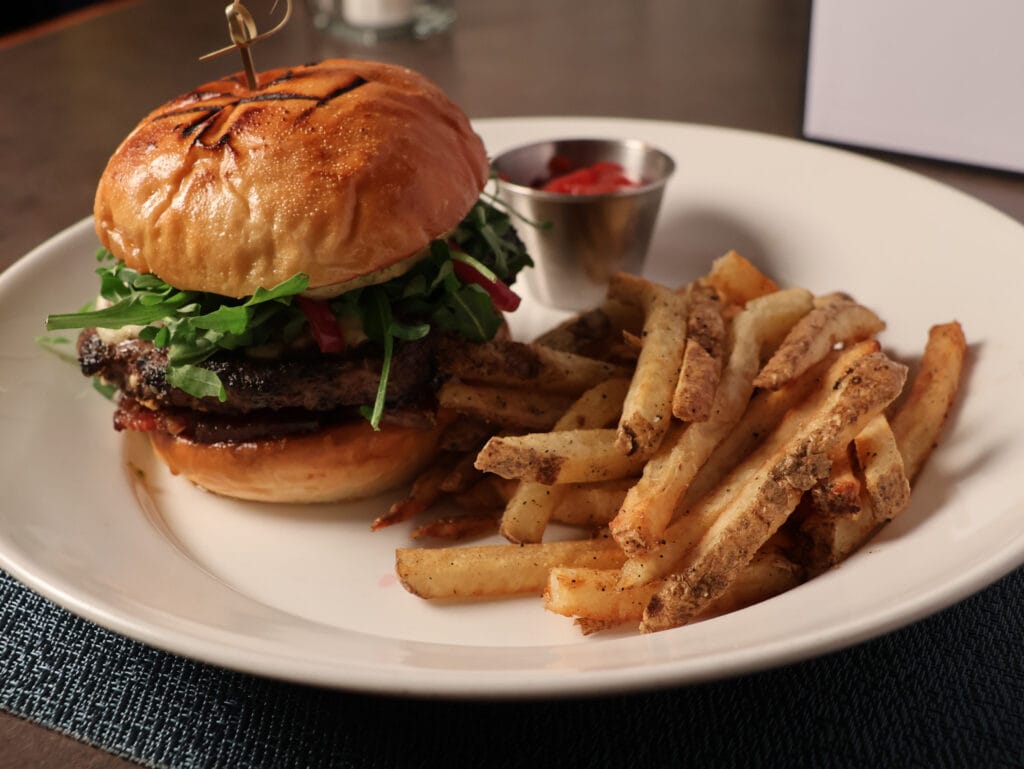 A grilled burger topped with lettuce, tomato, and onion is served on a toasted bun with a side of seasoned French fries and a small metal cup of ketchup on a white plate.