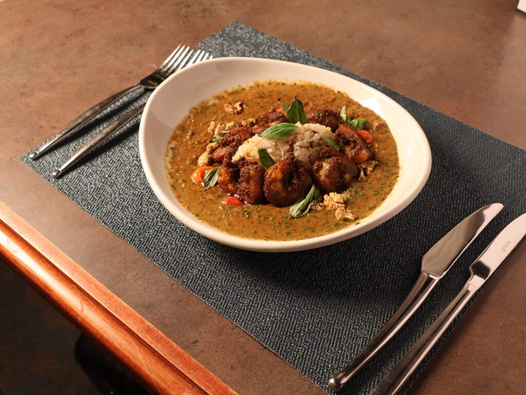 A white bowl of curry with rice, vegetables, and herbs is set on a dark placemat with a fork, knife, and spoon on a brown table.