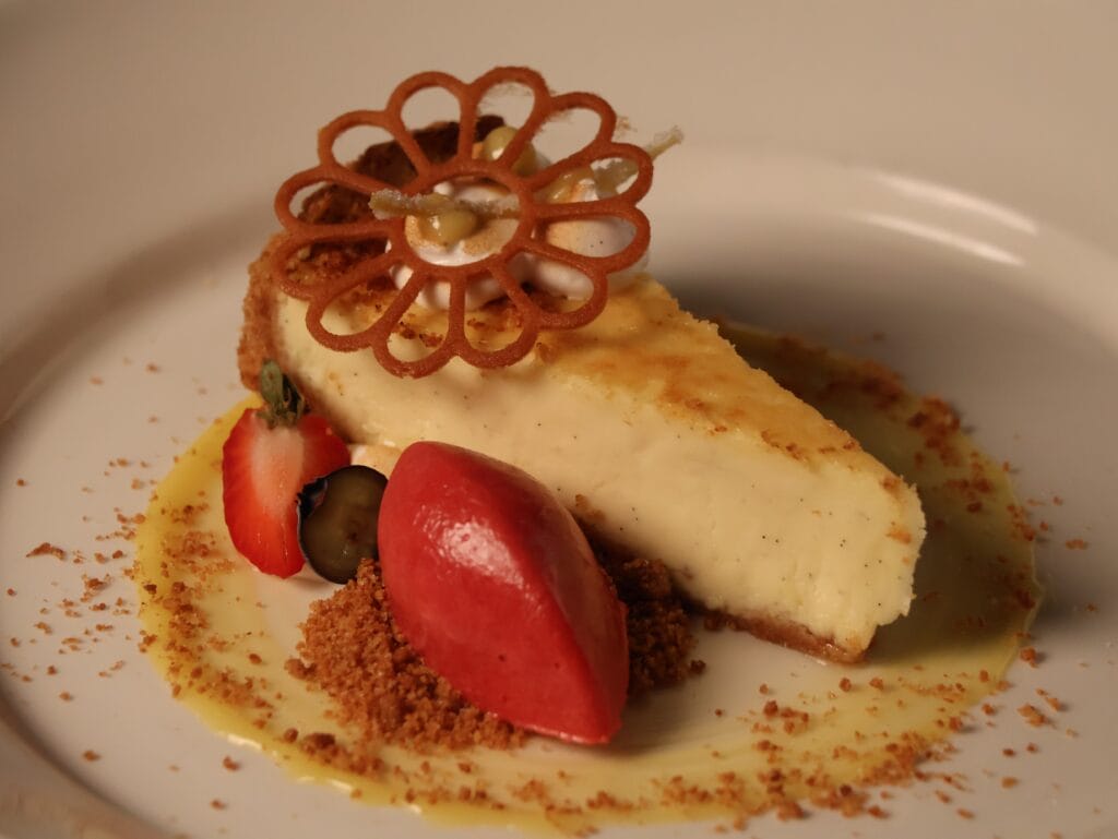 A slice of cheesecake garnished with a decorative cookie, strawberries, blueberries, crumbs, and a scoop of bright red sorbet, served on a white plate with a creamy sauce.