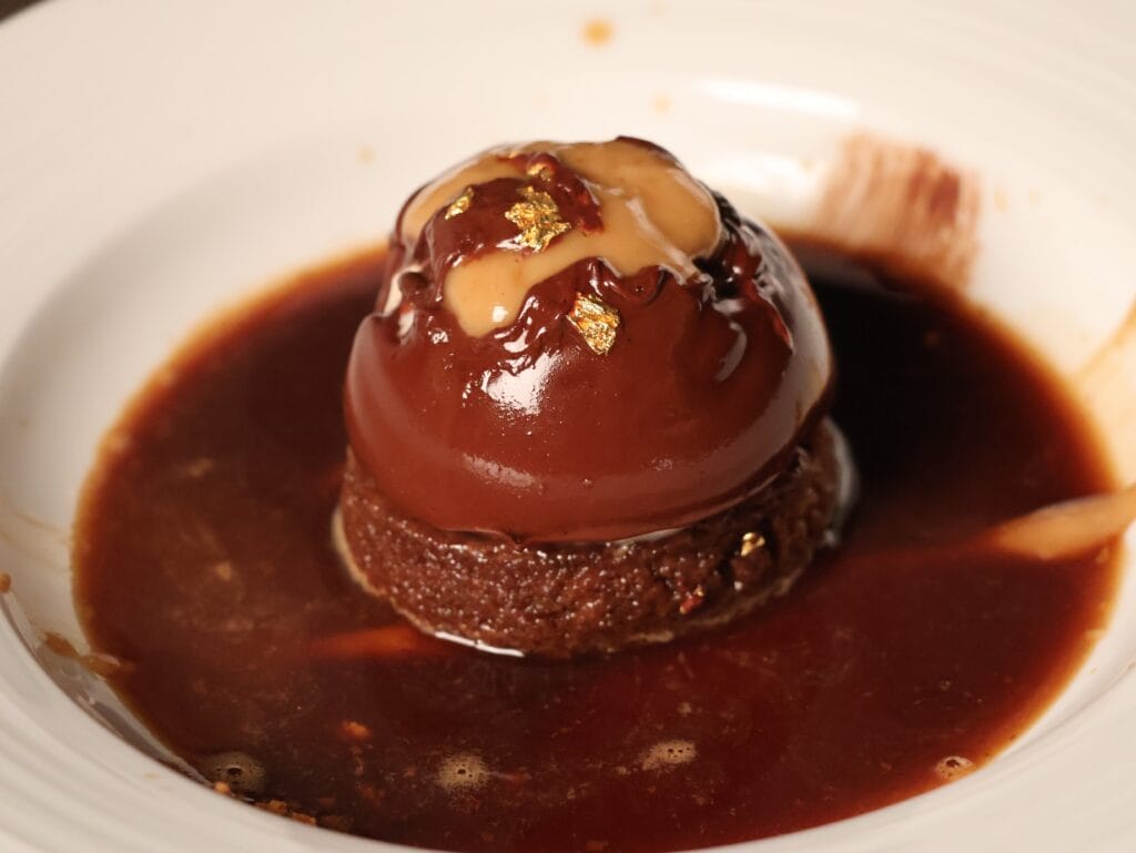 A glossy chocolate dome dessert with caramel and gold leaf accents sits on a round chocolate base, surrounded by a pool of rich chocolate sauce on a white plate.