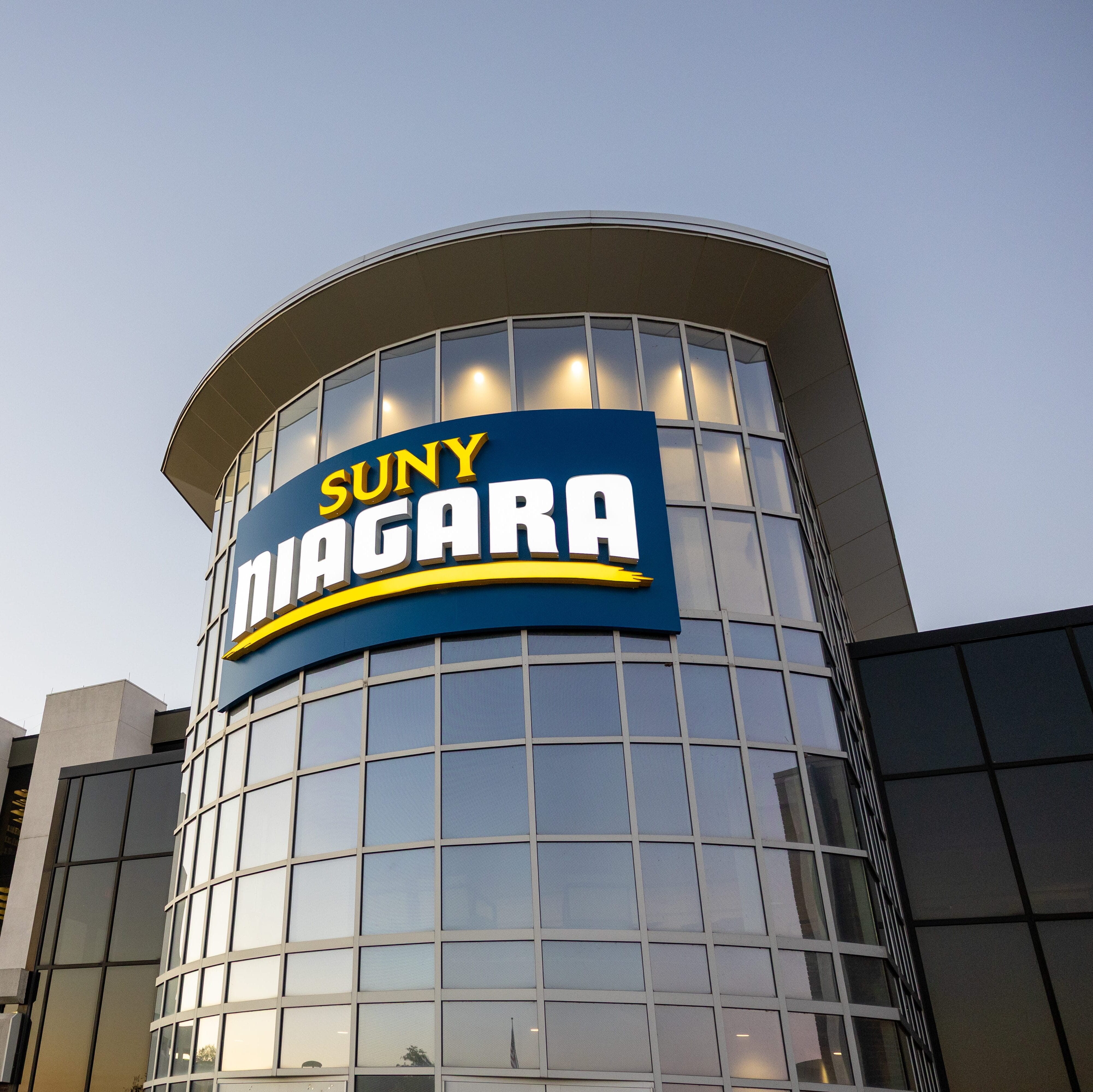 Modern building with a large curved glass facade displaying a bright sign that reads SUNY NIAGARA, with the sky clear in the background.