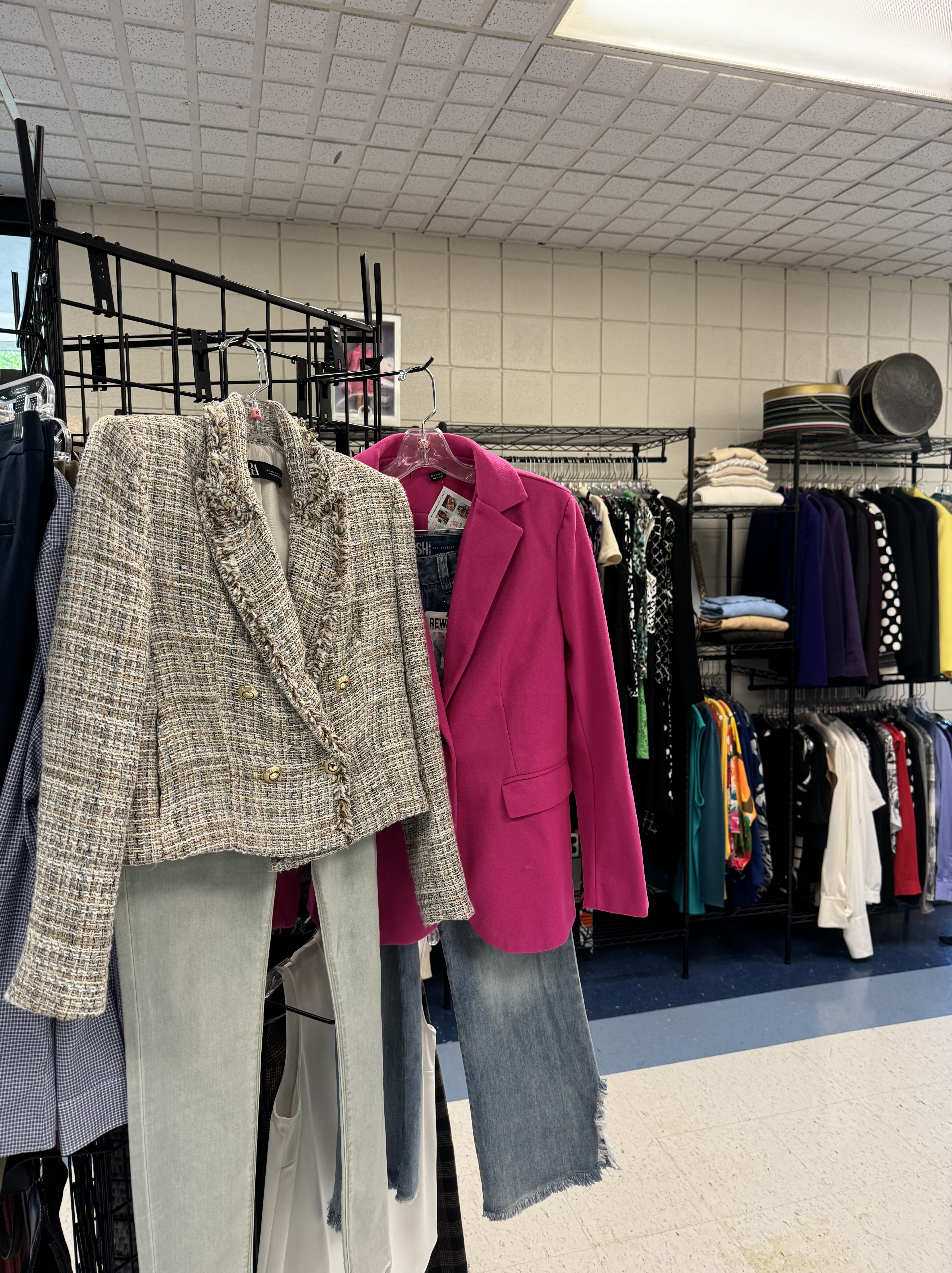Two outfits hang on display: one features a gray tweed blazer with light jeans, while the other has a bright pink blazer paired with frayed blue jeans. Clothing racks with various garments are visible in the background, showcasing options available through SUNY Niagara Student Basic Needs Resources.