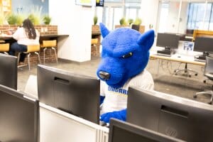 A person in a blue panther SUNY mascot costume sits at a computer in a library or study area, with other people working in the background.