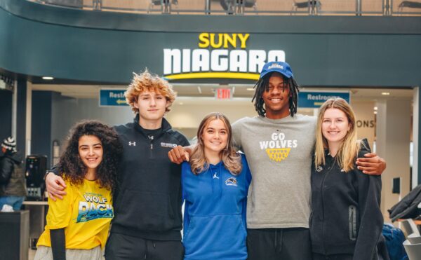 Five students, wearing SUNY Niagara and Wolves apparel, stand together smiling in a lobby area under a large SUNY Niagara sign.