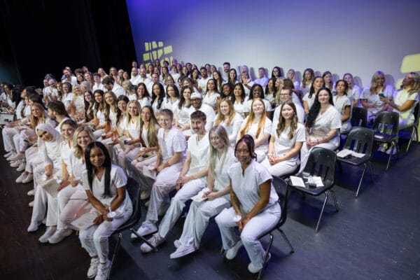 A large group of people dressed in white sit closely together on a stage, smiling at the camera. The background is plain, and the group appears to be at a formal event or ceremony.
