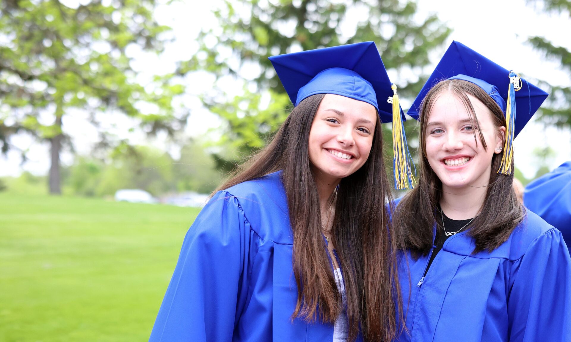 Two young women in blue graduation gowns and caps smile outdoors, standing close together on a grassy area, while another graduate adjusts their cap in the background.
