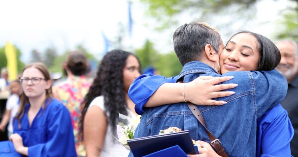 A graduate in a blue gown and cap hugs someone in denim. Other people, some in blue gowns, stand nearby. The scene is outdoors, and everyone appears joyful.