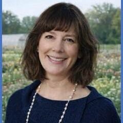 Woman with shoulder-length brown hair, wearing a navy blue top and a pearl necklace, smiling outdoors in front of a flower field and trees under a partly cloudy sky.