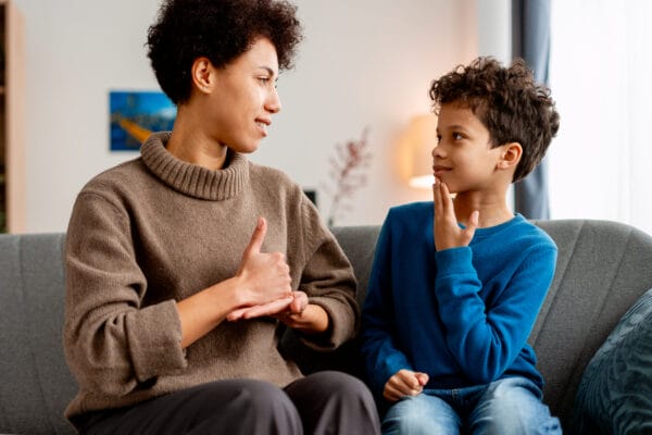 A woman and a child sit on a couch, facing each other and using sign language to communicate, both appearing focused and engaged in conversation.