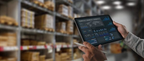 A person holds a digital tablet displaying data analytics dashboards in a warehouse with shelves stocked with boxes and products in the background.