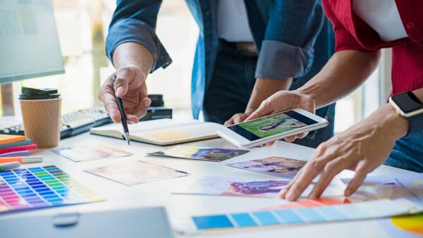 Two people collaborate at a desk with photos, a color swatch book, markers, a notebook, a coffee cup, and a tablet, suggesting a creative design or planning session.