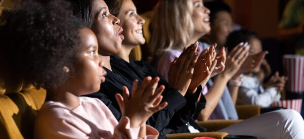 A group of people, including adults and children, sit in a theater, clapping and smiling as they watch a performance or movie.