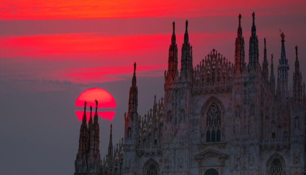 A red sunset behind the intricate Gothic spires of Milan Cathedral, casting a dramatic silhouette against the colorful sky.