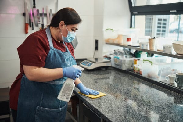 A person wearing a mask, gloves, and apron cleans a kitchen countertop with a spray bottle and cloth, with various utensils and containers visible in the background.