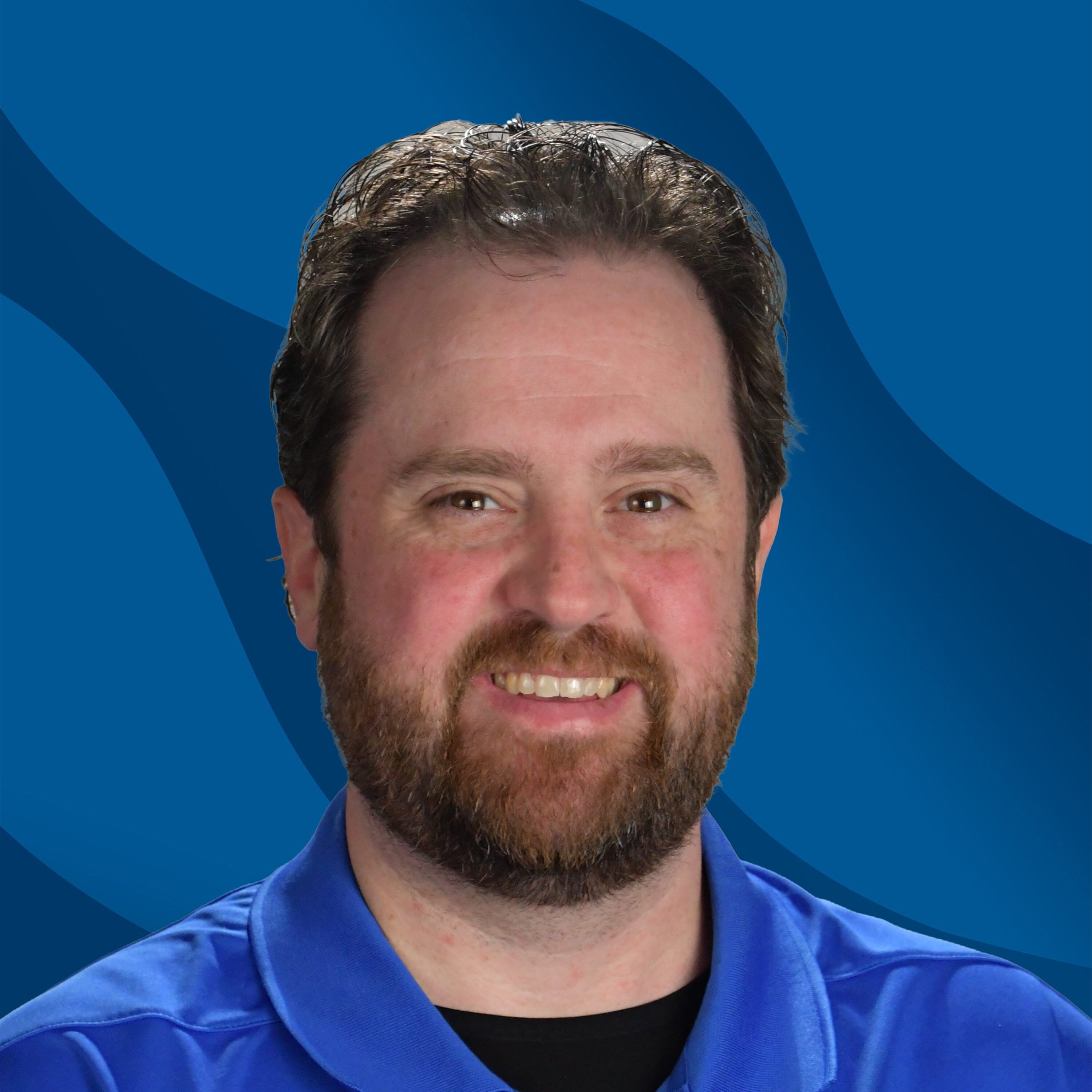 A man with brown hair and a beard smiles at the camera, wearing a blue collared shirt with a SUNY Niagara logo, in front of a blue wavy background.