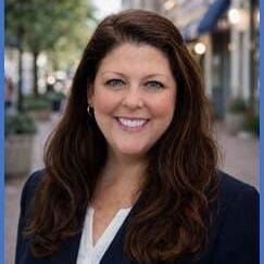 A woman with long brown hair wearing a navy blazer and white blouse smiles while standing on a city sidewalk with trees and storefronts in the background.