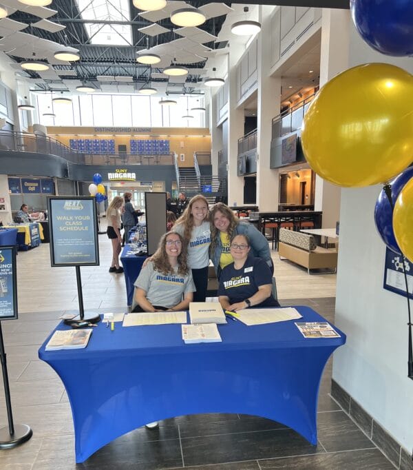 Four women stand and sit at a registration table with Niagara University signs inside a bright lobby. Two women sit, smiling, with two others standing behind them. Blue and yellow balloons and university banners are visible.