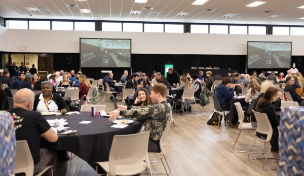 People sit at round tables in a large, bright event hall, eating and talking during the SUNY Niagara Cannabis Conference. Two large screens show a mirrored image of the room. The atmosphere seems relaxed and social as attendees mark their calendars to save the date for January 2025.